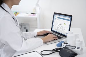 Doctor entering notes on a laptop amid medical devices — Ai scribing streamlines documentation, boosts physician productivity, improves patient satisfaction, and protects revenue..