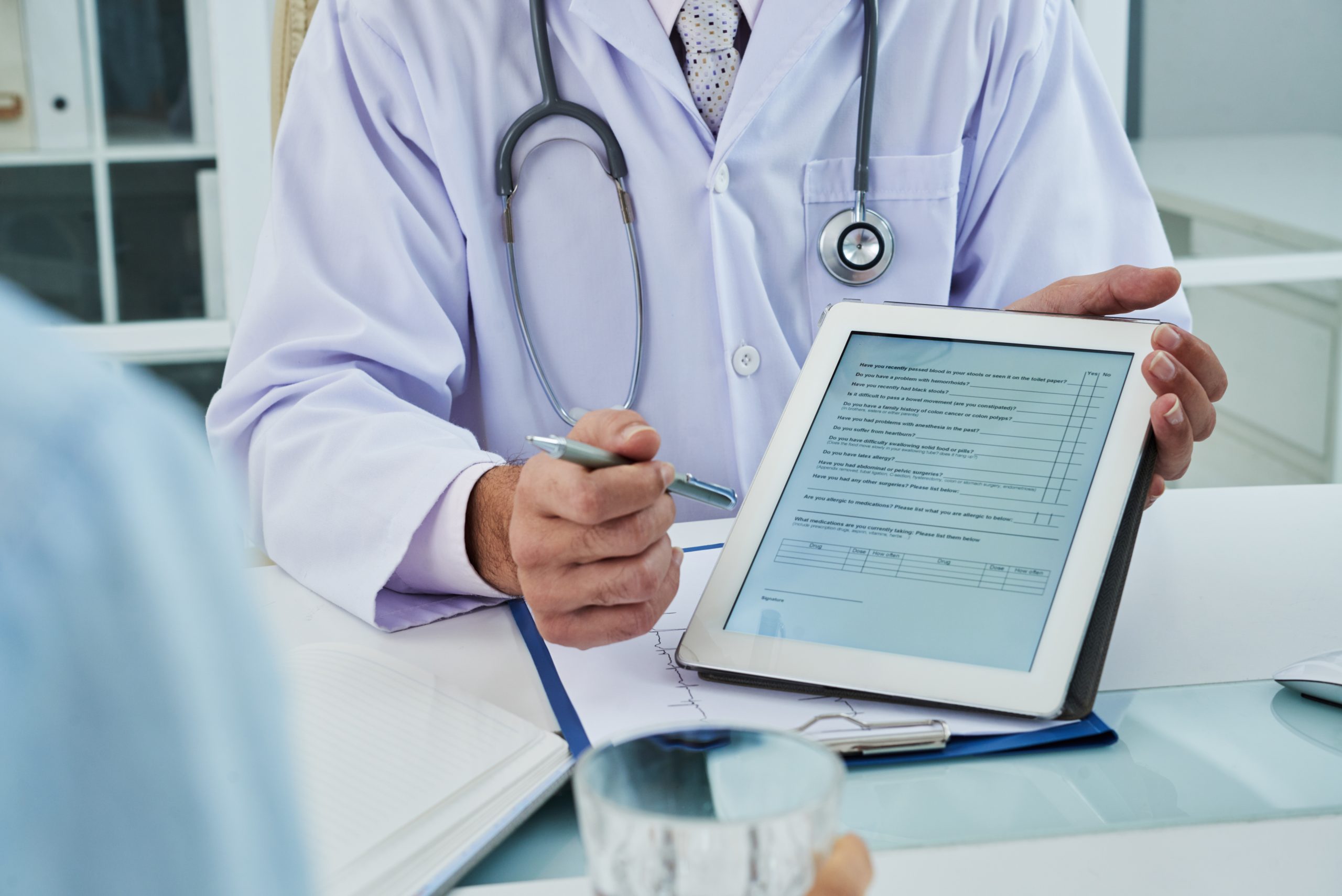 Doctor holding a tablet displaying a patient intake form during a consultation AI scribing capturing SOAP-note documentation electronically.