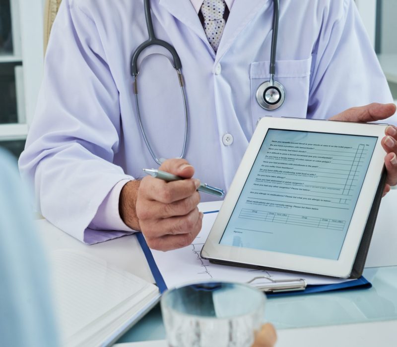 Doctor holding a tablet displaying a patient intake form during a consultation AI scribing capturing SOAP-note documentation electronically.